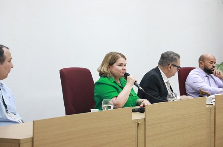 Fotografia de 4 pessoas, 3 homens e uma mulher. Ela está ao centro, segura um microfone e usa uma roupa verde, tem pele branca, 